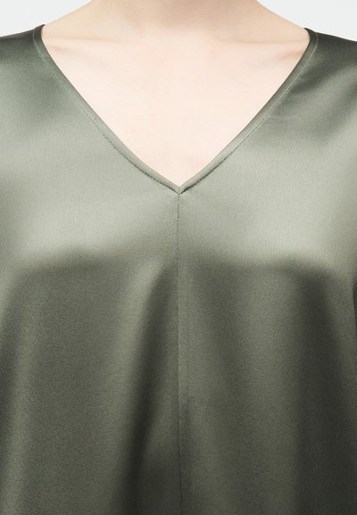 Κοντινό πλάνο ενός ατόμου που φοράει τοπ από σατέν σε ελιά πράσινο χρώμα με λαιμόκοψη σε σχήμα V, δείχνοντας την ομαλή υφή του υφάσματος και τις λεπτομέρειες της λαιμόκοψης.