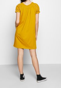 Abito giallo realizzato in tessuto morbido con maniche corte decorative. Presenta un collo arricciato e una vestibilità comoda. Indossato con sneaker alte nere.