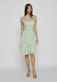 Φόρεμα σε μέντα πράσινη από chiffon με δαντελένιες λεπτομέρειες στους ώμους, φαρδύ σιγκράτημα και ζώνη που δένει μόνη της. Διαφανείς σάντες με τακούνι ολοκληρώνουν το σύνολο.