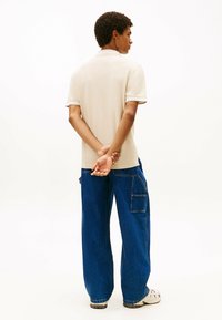 Junger Mann mit lockigem Haar, der ein beiges Poloshirt, lockere blaue Jeans mit Seitentasche und weiße Sneakers trägt, steht mit den Händen hinter dem Rücken.