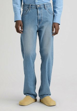 Osoba ubrana w jasnoniebieskie dżinsy, niebieską koszulę zapinaną na guziki oraz beżowe buty wsuwane stojąca na jednolitym szarym tle.