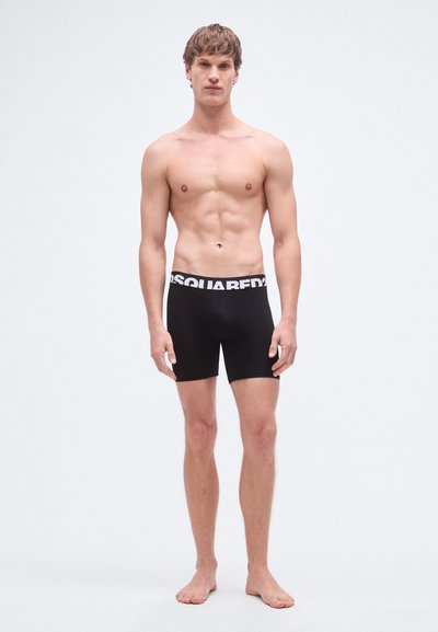 Črne bokser kratke hlače z belim logotipom na pasu, gladka tkanina, dolžina do sredine stegen in prilegajoč kroj, prikazane na stoječem moškem modelu.