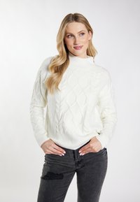 Blondynka w białym swetrze w sploty i podartych czarnych dżinsach, stojąca z rękami w kieszeniach na jednolitym tle.