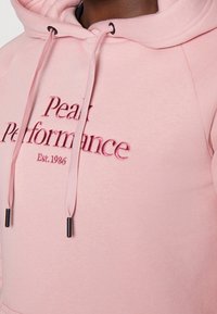 Felpa leggera rosa con ricamo del testo "Peak Performance", con cordini regolabili e un tessuto morbido e texturizzato. Presenta dettagli neri sulle punte dei cordini.