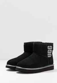 Czarne zamszowe botki za kostkę z okrągłym noskiem, srebrną podeszwą środkową oraz szarym logo "UGG" po boku. Miękka wyściółka dla komfortu.