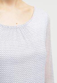 Kevyt, valkoinen pusero, jossa on hienovarainen tummansinisekuvio. Ominaisuuksiin kuuluu pyöreä kaula-aukko ja läpikuultavat pitkät hihat, sekä sileä ja hengittävä materiaali.