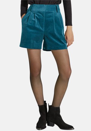 Shorts en velours côtelé bleu sarcelle taille haute portés avec des collants résille noirs et des bottines noires sur une personne portant un haut noir à manches longues.