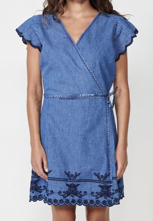 Kvinde iført en kortærmet denim slå-om-kjole med bølgede kanter og mørkt blomsterbroderi ved kanten.