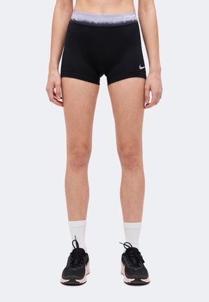 Kobieta ubrana w czarne spodenki Nike Pro, białe skarpetki i czarne buty sportowe stoi z ramionami luźno opuszczonymi wzdłuż ciała.