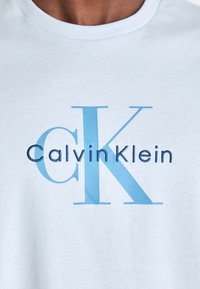 Camiseta de algodón azul claro con un audaz logo bordado de "Calvin Klein" en azul más oscuro y una gran "K" en un tono azul más claro.