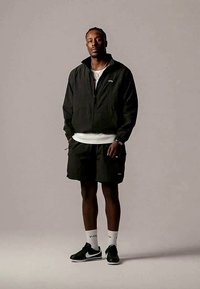 Crna jakna na patentni zatvarač preko bijele košulje, u kombinaciji s crnim kratkim hlačama. Nosite crne tenisice s bijelim detaljima i bijele čarape. Jednostavna pozadina.