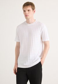 Tricou alb, cu mânecă scurtă, având un model texturat în grilă, guler rotund și o croială confortabilă. Asortat cu pantaloni negri.