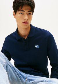 Camisa polo de color navy con tejido texturizado y cuello, que presenta un pequeño parche de logo en el pecho. Se lleva con unos vaqueros azules desgastados.