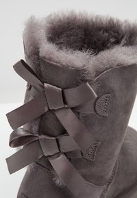 Grå ruskindsstøvle med en blød, fuzzy foring og dekorative mørke satinfwister på bagsiden, suppleret med subtile syninger.