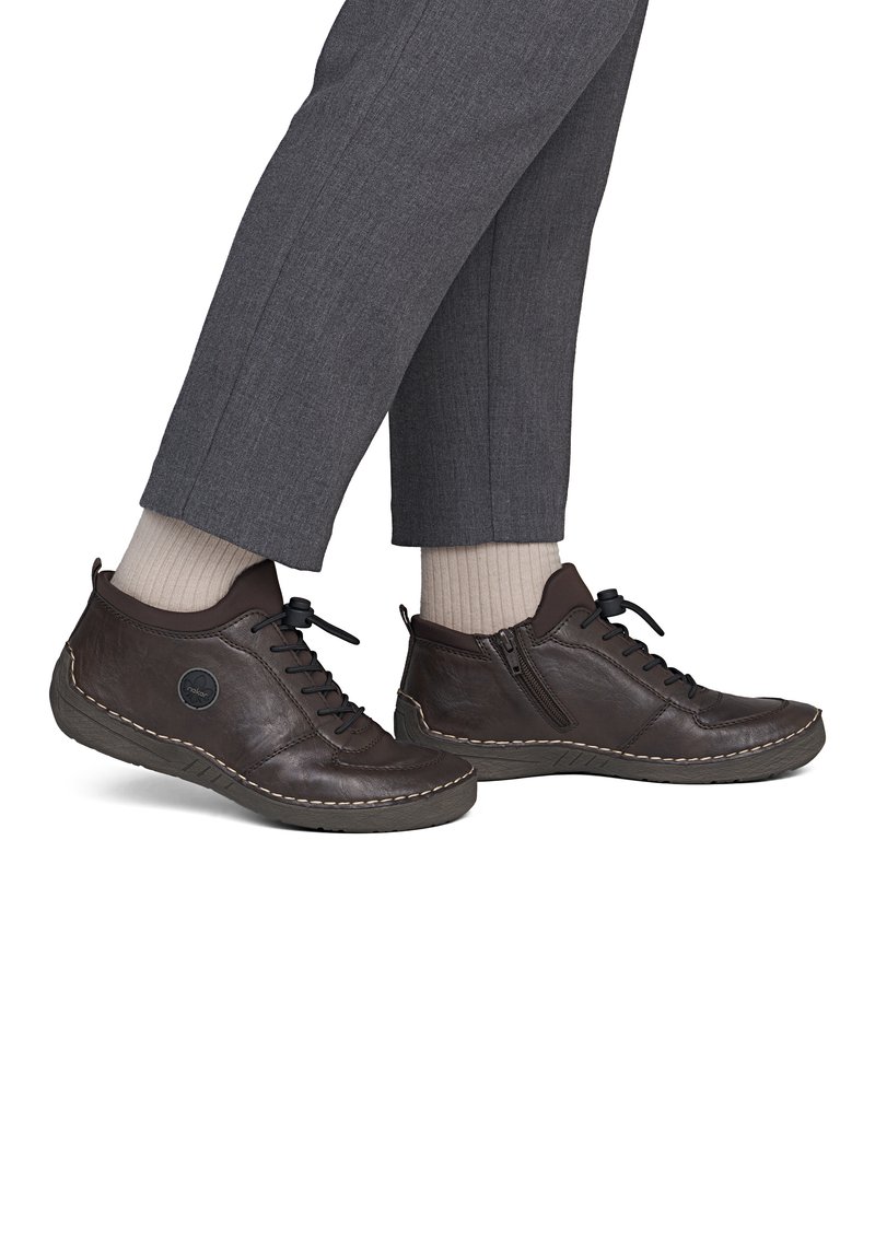 Chaussures décontractées en cuir marron avec lacets et fermetures éclair latérales, portées avec des chaussettes beiges et un pantalon gris, vues de côté sur fond blanc.