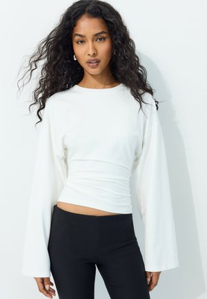 Femme aux longs cheveux bouclés portant un haut blanc ajusté à manches longues et un pantalon noir, debout sur un fond blanc.