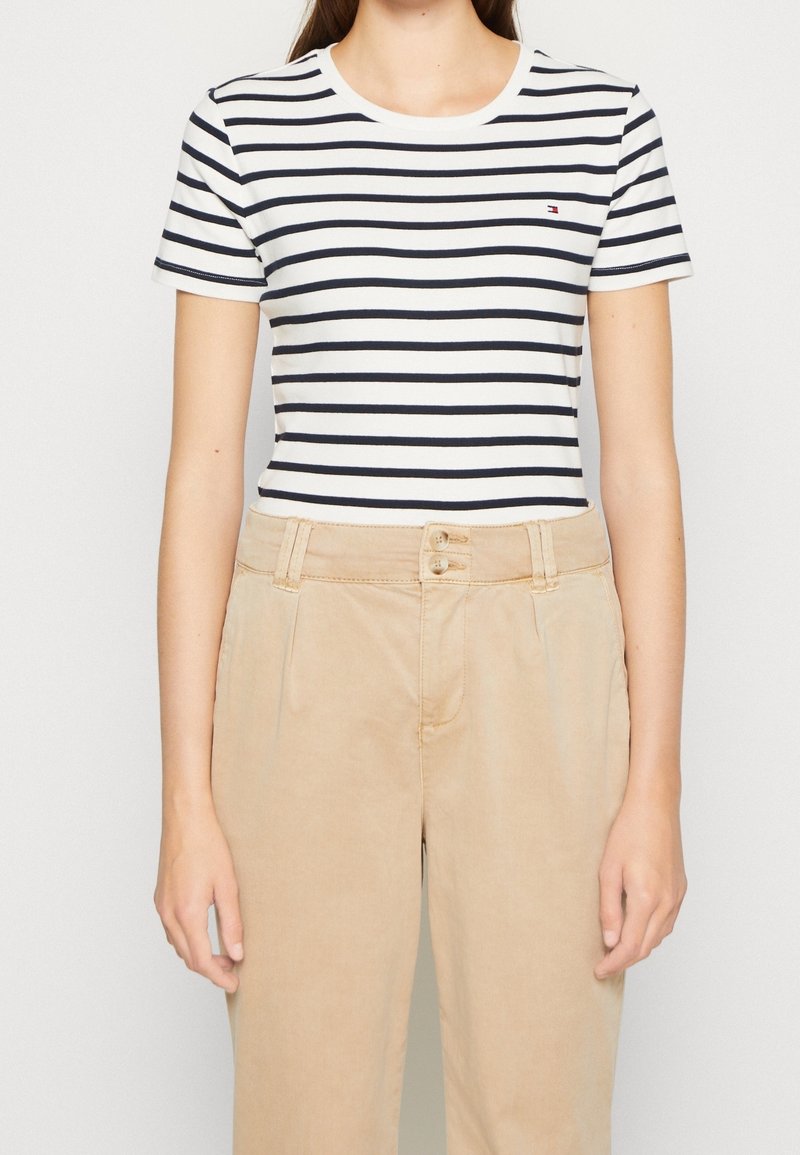 T-shirt blanc avec des rayures horizontales bleues marines, décolleté rond, associé à un pantalon beige taille haute texturé avec une fermeture à boutons.