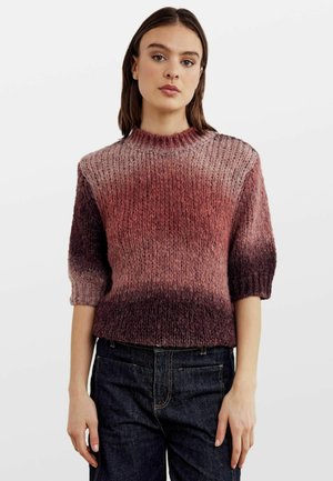 Kobieta ubrana w sweter z gradientowym wzorem w odcieniach różu i burgunda, z krótkimi rękawami, w połączeniu z ciemnoniebieskimi dżinsami, stojąca na jednolitym tle.