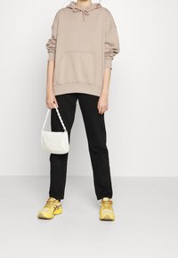 Persona che indossa un hoodie beige, pantaloni neri, sneakers gialle, mentre tiene una piccola borsa bianca con una tracolla a catena su uno sfondo bianco uniforme.