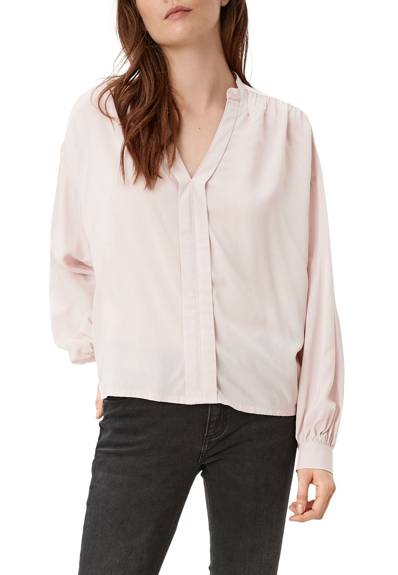 Blouse rose clair avec un col en V, manches longues, et détails froncés aux épaules. Présente un ourlet droit et un léger éclat.