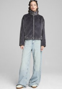 Giacca zip-up in pelliccia sintetica grigia con alto colletto, vestibilità ampia e logo ricamato. Abbinata a jeans in denim azzurri a gamba larga e sneakers bianche.