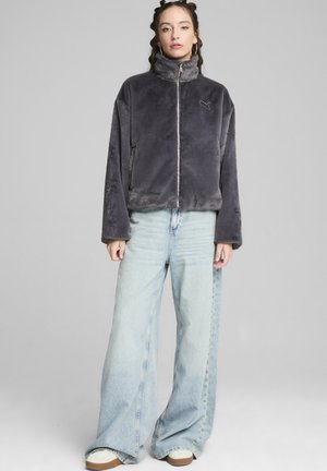 Giacca zip-up in pelliccia sintetica grigia con alto colletto, vestibilità ampia e logo ricamato. Abbinata a jeans in denim azzurri a gamba larga e sneakers bianche.