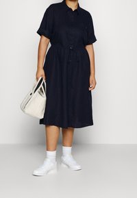 Robe bleu marine à boutons, à manches courtes retroussées, avec une taille à cordon, associée à des sneakers blanches et un sac crème avec des accents noirs.