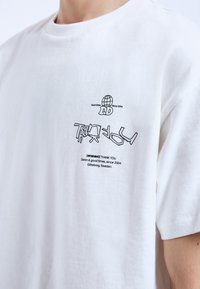 Bílé bavlněné tričko s černým grafickým textem a vzory na přední straně, s kulatým výstřihem a krátkými rukávy.