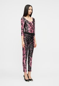 Γυναίκα που στέκεται φορώντας στενή ολόσωμη φόρμα με μαύρο και ροζ σχέδιο, μανίκια μέχρι τον αγκώνα και μαύρες μυτερές γόβες, μπροστά σε απλό φόντο.