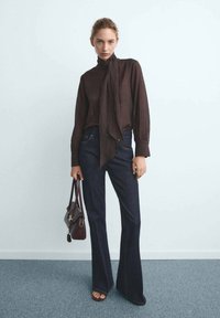 Eine Frau trägt eine dunkelbraune Bluse mit langen Ärmeln und einem Halsband, dunkle blaue Schlagjeans, braune Sandalen mit offenen Zehen und hält eine dunkelbraune Handtasche.