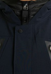 Granatowa kurtka z kapturem, zapięciem na zamek błyskawiczny i zatrzaskiem oraz teksturowaną tkaniną, z dwoma kieszeniami na zamki z przodu.