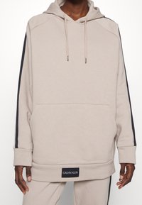 Sweatshirt à capuche beige avec des manches longues, poche avant et rayures noires sur les côtés arborant la marque "CALVIN KLEIN". Texture de tissu douce.