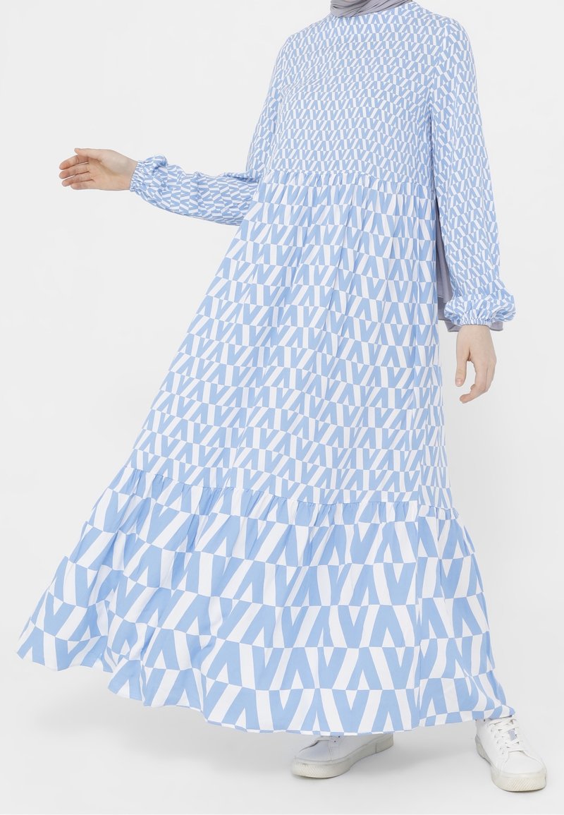 Femme portant une robe longue à manches longues bleu et blanc à motifs, avec un ourlet volanté et des baskets blanches, debout sur fond blanc.