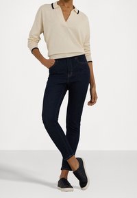 Žena v béžovém pleteném svetru s dlouhým rukávem, tmavě modrých skinny džínách a černých teniskách, stojící s jednou rukou v kapse na bílém pozadí.