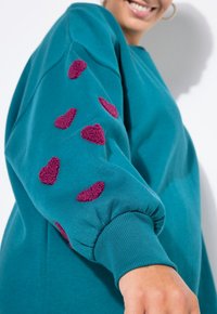 Sudadera azul verdoso con puños elásticos, decorada con apliques de peluche en forma de corazón burdeos en las mangas. Tejido suave con un corte holgado.