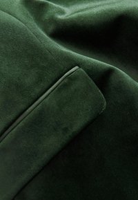 Dunkelgrüner Samtstoff mit einer glatten Textur, der eine subtile Glanzwirkung aufweist und eine schräge Taschenverzierung in der oberen rechten Ecke hat.