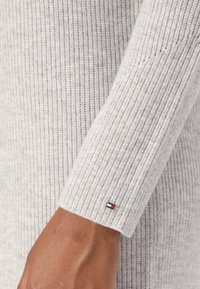 Manche de pull gris côtelé avec un tricot texturé. Comprend un petit logo de marque en rouge, blanc et bleu sur le poignet.