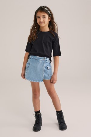 Maglietta nera, gonna di jeans blu con tasche frontali e stivaletti neri alla caviglia. L'outfit presenta uno stile casual a strati con una vestibilità confortevole.