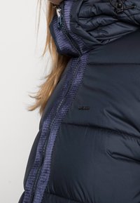 Marinblå puffjacka med dragkedja, texturerade blå detaljer längs dragkedjan och en vadderad design med hög krage.