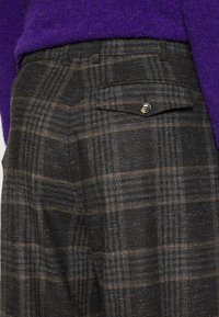 Pantaloni în carouri de culoare gri închis cu dungi deschise și maronii. Au un buzunar posterior cu închidere prin buton. Fabricat dintr-un material texturat.