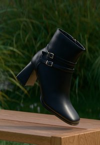 Μαύρη γόβα αστραφτερής υφής με δύο πλάγιες αγκράφες, τετράγωνη μύτη και μέτριο φαρδύ τακούνι. Τοποθετημένη σε φυσικό φόντο.