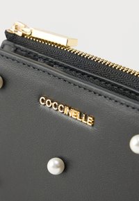 Musta nahkainen lompakko, jossa on kullanvärinen vetoketju. Etupuolella koristeina valkoiset tekokuplathelmet ja kullanvärinen "Coccinelle"-logo.