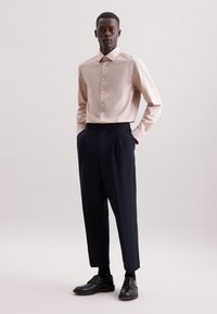 Camicia button-up rosa chiaro in tessuto morbido, abbinata a pantaloni pieghettati blu scuro e scarpe nere, il tutto su uno sfondo neutro.
