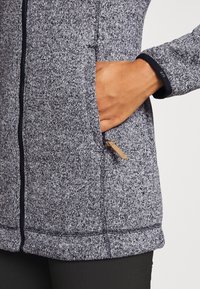 Giacca a zip grigia con texture, rifinita con un bordo nero, che presenta una tasca laterale con una zip in pelle e dettagli di cucitura visibili.