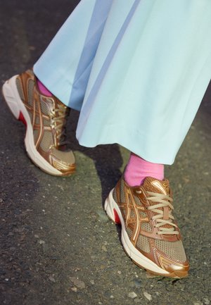 Brune og gyldne mesh-sneakers båret med lyserøde sokker og lyseblå vide bukser på asfaltbelægning.