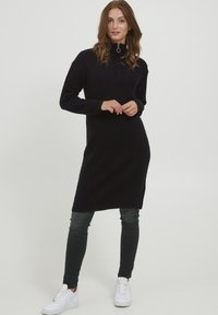 Abito in maglia blu navy con colletto alto, caratterizzato da una mezza cerniera al collo. Abbinato a jeans scuri e sneaker bianche.