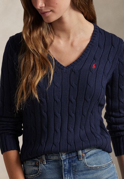 Mujer vistiendo un suéter azul marino de punto trenzado con cuello en V y un pequeño logo rojo bordado, combinado con vaqueros azules.