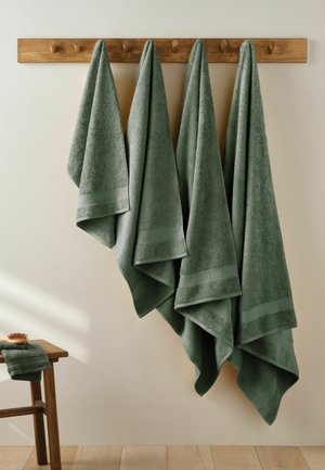 Vier groene handdoeken hangen aan houten haken boven een houten bankje met een opgevouwen handdoek en een borstel erop.