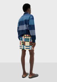 Suéter a rayas azul y gris, pantalones cortos de patrón multicolor con rectángulos y líneas, y zapatos grises sin cordones. Vista desde atrás.