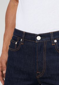 Muž nosící tmavě modré džíny s oranžovým šitím a bílým knoflíkem, v kombinaci s bílou košilí zastrčenou dovnitř.